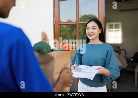 Asiatico consegna uomo che tiene la penna e maneggia il sacchetto di cibo, frutta e verdura dare alla cliente femminile di fronte alla casa Foto Stock