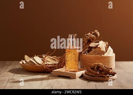 Una bottiglia di siero in vetro riempita di essenza gialla in piedi su un podio. La radice di Angelica e il Bai Zhu sono contenuti in alcuni oggetti di legno Foto Stock