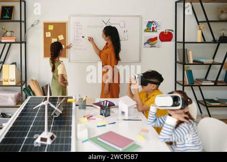 I bambini con occhiali per realtà virtuale guardano un video educativo quando un compagno di classe parla con l'insegnante Foto Stock