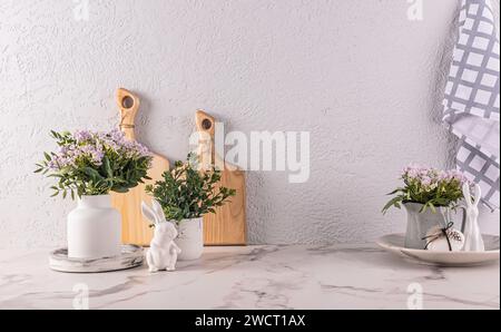 Fiori primaverili in vaso e brocca sul ripiano della cucina con decorazioni pasquali. taglieri, asciugamani. Statuette coniglietto pasquale Foto Stock