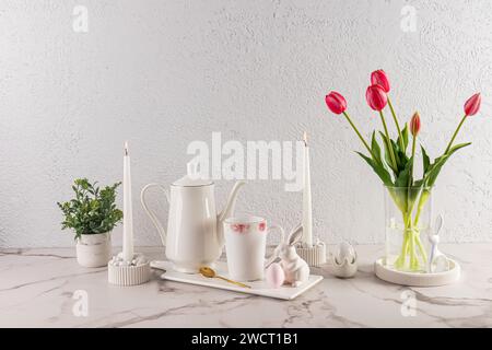 Natura morta elegante per le vacanze di Pasqua. Caffettiera bianca su vassoio bianco in ceramica con tazza e personaggi coniglietto pasquale. vaso di fiori e candele Foto Stock