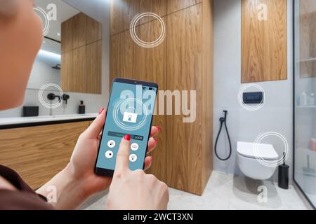 Primo piano smartphone in mano femminile con interfaccia app Smart Home System sullo schermo del cellulare, giovane donna che controlla tutto il sistema di casa tramite telefono Foto Stock