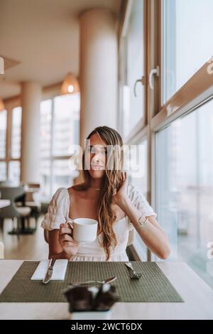 Una donna attraente che si gode un momento vicino alla finestra, assaporando una tazza di caffè appena fatto Foto Stock