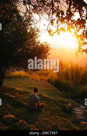 Una persona siede in una tranquilla meditazione su una collina erbosa mentre il caldo bagliore del tramonto bagna il paesaggio in sfumature dorate, creando una scena tranquilla Foto Stock
