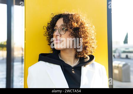 Una giovane donna premurosa con un cappotto bianco e occhiali si appoggia su una parete gialla, guardando in lontananza Foto Stock
