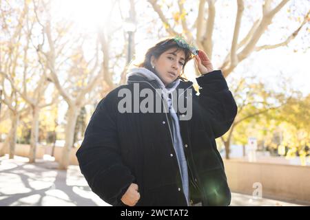 Una giovane donna con una giacca nera e occhiali da sole si trova in un parco con alberi autunnali, crogiolati alla luce del sole Foto Stock