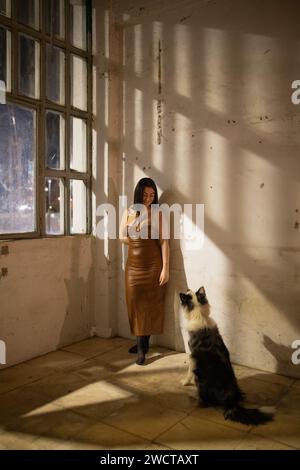 In una stanza leggermente illuminata, una donna sta guardando lontano con un grande cane seduto con attenzione al suo fianco Foto Stock
