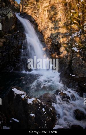 Una tranquilla cascata scende lungo un terreno accidentato circondato da foreste, con rocce innevate che circondano la scena Foto Stock