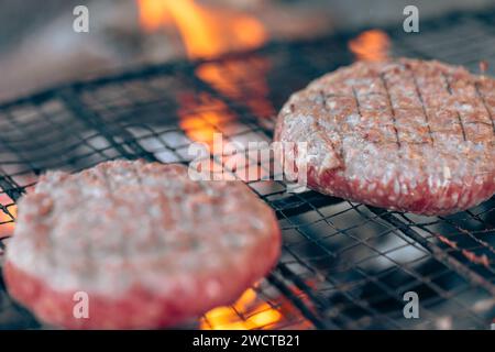 Primo piano di due hamburger che cucinano su una griglia con fiamme sullo sfondo, catturando l'essenza di un barbecue Foto Stock