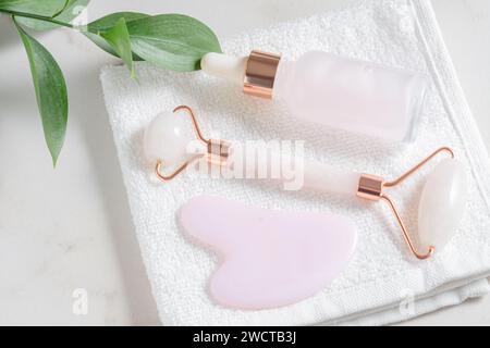 Rullo facciale al quarzo rosa, strumento gua Sha, flacone per siero e foglia verde fresca su un asciugamano bianco testurizzato per la cura della pelle Foto Stock