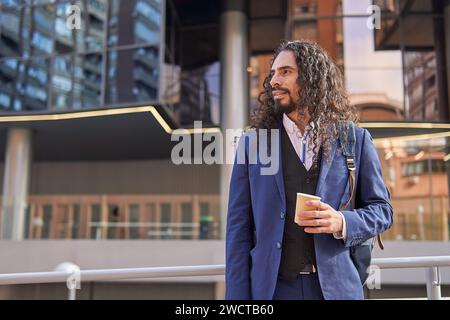 Dal basso sorridente uomo d'affari latino maturo con i capelli lunghi che reggono una tazza di caffè, in piedi vicino a un moderno edificio urbano Foto Stock