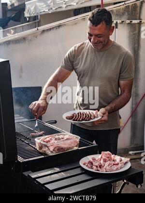 Un maschio adulto allegro e concentrato che guarda in basso mentre si è in piedi vicino a grigliate affumicanti con salsicce e carne grigliata sul piatto insieme a una scatola di vetro Foto Stock