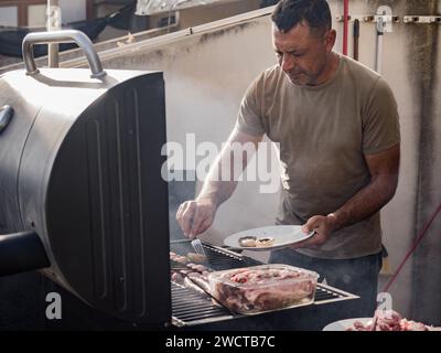 Maschio adulto concentrato che guarda in basso mentre si è in piedi vicino a griglia per barbecue fumatori con funghi sul piatto mentre si grigia carne e salsicce nel tetto ope Foto Stock