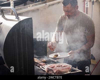 Maschio adulto concentrato che guarda in basso mentre si è in piedi vicino a griglia per barbecue fumatori con funghi sul piatto mentre si grigia carne e salsicce nel tetto ope Foto Stock