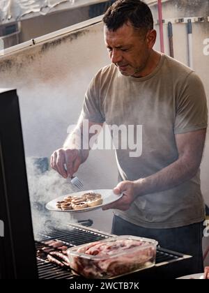 Maschio adulto concentrato che guarda in basso mentre si è in piedi vicino a griglia per barbecue fumatori con funghi sul piatto mentre si grigia carne e salsicce nel tetto ope Foto Stock