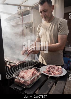 Maschio adulto concentrato che guarda in basso mentre si è in piedi vicino a griglia per barbecue affumicato con salsicce e carne grigliata sul piatto e contenitore in vetro Foto Stock