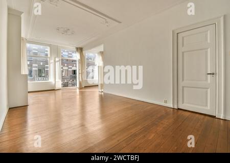 Un elegante e vuoto soggiorno caratterizzato da pavimenti in legno duro lucido, grandi finestre con tende e intricate modanature del soffitto. Foto Stock