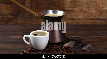 Caffè turco. Cezve e tazza con bevanda appena preparata, chicchi di caffè e cioccolato su un tavolo di legno. Design banner Foto Stock