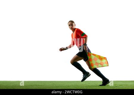 Assistente arbitro, giovane donna in uniforme in movimento sul campo, corsa con la bandiera, guida il controllo sul gioco sullo sfondo bianco dello studio Foto Stock