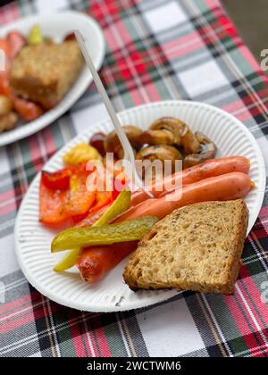 Un pasto informale all'aperto con salsicce alla griglia, verdure e pane su un piatto di carta. Foto Stock