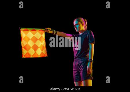 Giovane donna, assistente arbitro che alza la bandiera, il che significa posizione in fuorigioco sullo sfondo nero dello studio in luce al neon Foto Stock