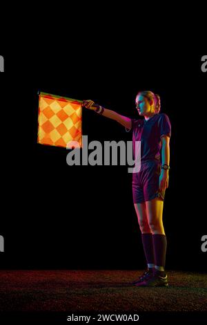 Giovane donna, assistente arbitro che alza la bandiera, il che significa posizione in fuorigioco sullo sfondo nero dello studio in luce al neon Foto Stock