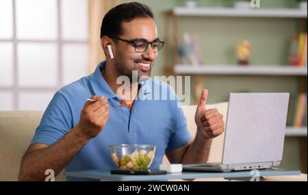 Uomo indiano felice che mangia Macedonia di frutta mostrando pollice su su su su su videochiamata online a casa - concetto di soddisfazione, comunicazione e alimentazione sana. Foto Stock