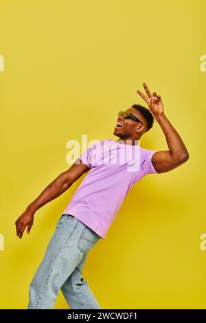 allegro ragazzo afroamericano con occhiali da sole e maglietta viola con il simbolo della vittoria in giallo Foto Stock