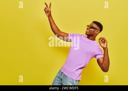uomo afro-americano sorridente con eleganti occhiali da sole e maglietta viola con il simbolo della vittoria in giallo Foto Stock
