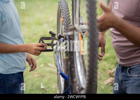 Primo piano del padre indiano con il figlio che ripara o controlla la bicicletta al parco durante la passeggiata mattutina - concetto di insieme, manutenzione e genitori Foto Stock