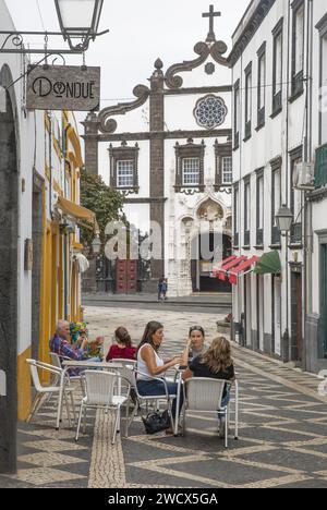 Portogallo, arcipelago delle Azzorre, isola di Sao Miguel, Ponta Delgada, persone sulla terrazza di un caffè in un vicolo con ciottoli bianchi e neri, di fronte a una chiesa barocca Foto Stock
