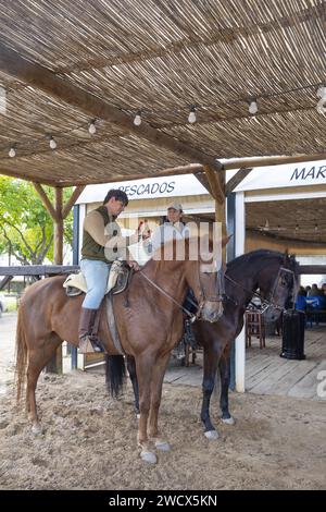 Spagna, Andalusia, El Rocío, coppia di cavalieri a cavallo e birre in mano con bicchieri al bancone esterno a cavallo di un bar in una strada sabbiosa Foto Stock