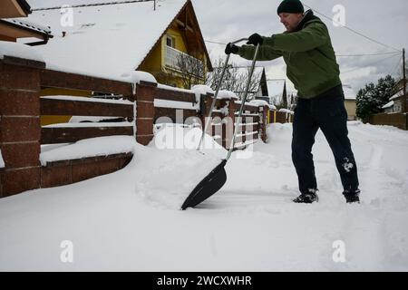 un ragazzo con giacca, cappello e guanti scuote la neve davanti alla casa dopo la bufera di neve - vista ravvicinata. Foto Stock