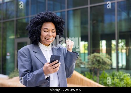 Foto ravvicinata di una giovane donna afroamericana in tuta in piedi vicino al centro degli uffici, che tiene e guarda il telefono cellulare, felice di mostrare il gesto della vittoria con la mano. Foto Stock