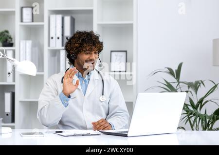 Coinvolge un medico indù maschio in un cappotto bianco con cuffie che salutano la fotocamera, fornendo una calda consulenza virtuale dal suo ufficio ben illuminato. Foto Stock