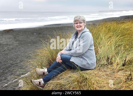 Una donna matura siede sulla costa dell'Oregon all'alba. Indossa una felpa grigia con cappuccio, jeans e scarpe da tennis. Foto Stock