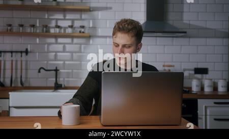 Un giovane biondo siede in cucina con una tazza di caffè, controlla la posta, i social media o le notizie sul portatile, beve il caffè Foto Stock