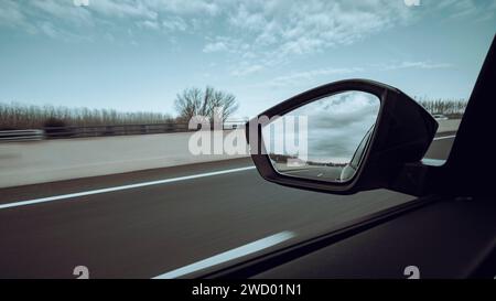 Vista sul finestrino sinistro di un'auto stradale su un'autostrada, su un bellissimo paesaggio boschivo con uno specchio retrostante Foto Stock