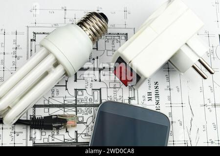 Lampada a risparmio energetico, smartphone e spina di rete sui disegni di costruzione, immagine simbolica per la casa intelligente Foto Stock