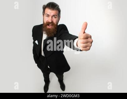 Emozionato uomo d'affari stupito in vestito nero sorridente e mostrando pollici in su. Fuga dalla crisi. Faccia divertente. Ottima idea. Ritratto di uomo eccitato gesturing Foto Stock