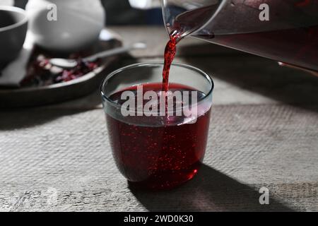 Versare il tè aromatico all'ibisco dalla teiera al vetro su un tavolo di legno, primo piano Foto Stock