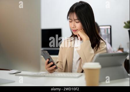 Una donna d'affari asiatica felice o un'assistente di marketing che utilizza il suo smartphone alla sua scrivania in ufficio. controllo dei messaggi, lettura dei testi, scorrimento Foto Stock