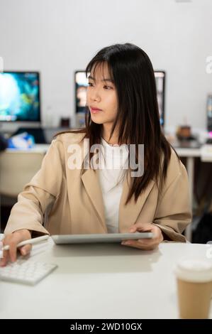 Una donna d'affari asiatica bella e concentrata, o una dipendente d'ufficio, lavora alla sua scrivania in ufficio, lavorando al computer. Foto Stock