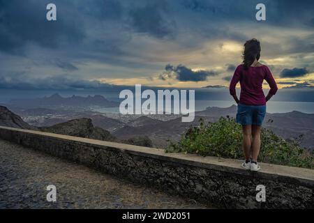 Una ragazza contemplativa si trova in un punto panoramico che si affaccia sulla città di Mindelo, São Vicente, Capo Verde, con uno skyline spettacolare sagomato contro una dinna Foto Stock