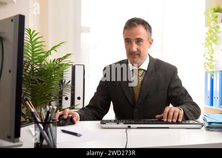 Uomo d'affari con abito grigio e cravatta dorata che lavora con un computer che digita e usa il mouse in ufficio guardando un monitor. Foto Stock