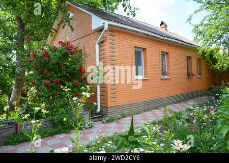 Bella casa in mattoni con tetto in amianto, grondaia, vialetto in un intimo giardino con alberi e rose Foto Stock