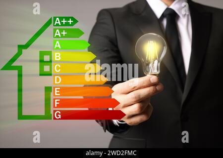 Classe di efficienza energetica e uomo che tiene una lampadina illuminante su sfondo grigio, primo piano Foto Stock