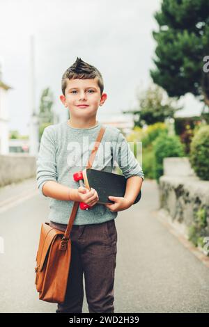 Ritratto all'aperto di un simpatico ragazzino che indossa una borsa in pelle marrone a spalla e tiene in mano lo skateboard. Concetto di ritorno a scuola. Immagine tonificata dell'aspetto della pellicola Foto Stock