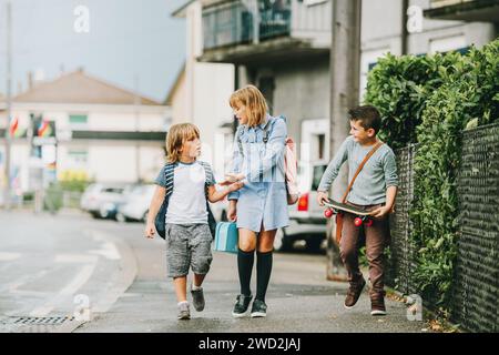 Gruppo di 3 divertenti scolari che tornano a scuola insieme, indossando zaini, tenendo in mano il pranzo al sacco e lo skateboard Foto Stock