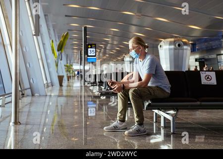 Uomo con power bank in aeroporto, ricarica il telefono per rimanere collegato durante il viaggio, prima del volo all'area di partenza. Passeggero con maschera facciale Foto Stock